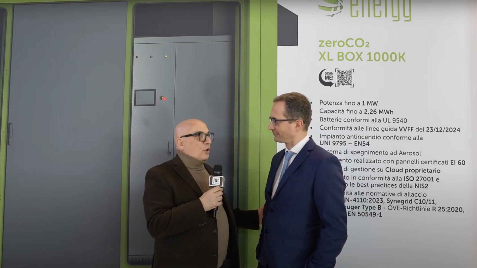 Due uomini in giacca parlano durante un’intervista davanti allo stand Energy e al pannello XL BOX 1000K
